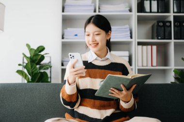 Çekici, mutlu Asyalı öğrenci üniversite kütüphanesinde okuyor, kanepede oturuyor, akıllı telefon kullanıyor.