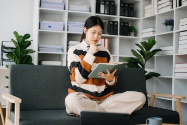 Kendine güvenen Asyalı kadın oturma odasında kitap okuyor ev ofisinin kanepesinde.