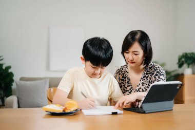 Evde eğitim gören Asyalı küçük çocuk internetten öğreniyor ve annesiyle tablet kullanarak ödevlerini yapıyor..