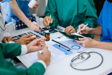 Tıbbi ekip, beyaz laboratuvar önlüklü ve ameliyat önlüklü doktorlarla bir toplantı yapıyor. Masada oturmuş, tıp endüstrisinde tablet kullanan hastaları tartışıyorlar.