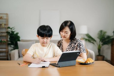 Evde eğitim gören Asyalı küçük çocuk internetten öğreniyor ve annesiyle tablet kullanarak ödevlerini yapıyor..