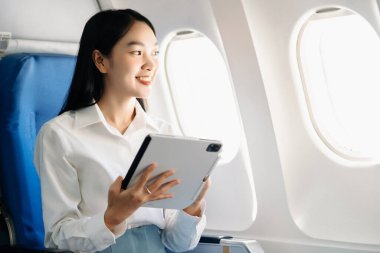 Çekici Asyalı kadın uçak yolcusu kablosuz bağlantıyla çalışırken rahat koltukta oturuyor. Şık bir şekilde seyahat et, sabahları zarafetle çalış.