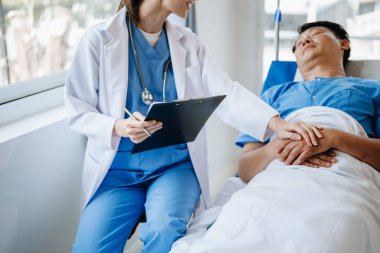 Kadın doktor, hastanede ya da klinikte salin solüsyonu alan erkek hasta elini yatakta tutuyor.