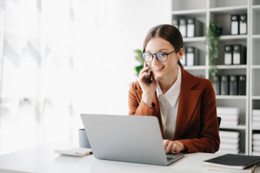 Beyaz iş kadını telefonda konuşuyor ve ofiste otururken gülümseyen bir dizüstü bilgisayar kullanıyor.