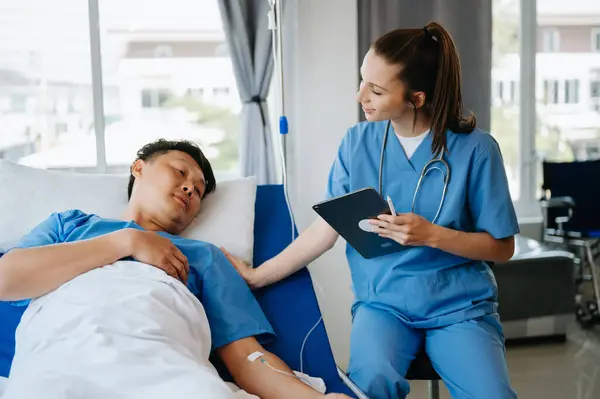 Friendly Female Head Nurse Making Rounds does Checkup on Patient Resting in Bed. She Checks tablet while Man Fully Recovering after Successful Surgery in modern hospital