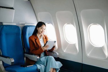 Çekici Asyalı kadın uçak yolcusu kablosuz bağlantıyı kullanarak dijital tablet üzerinde çalışırken rahat koltukta oturuyor. Stil içinde seyahat et, zarafetle çalış.