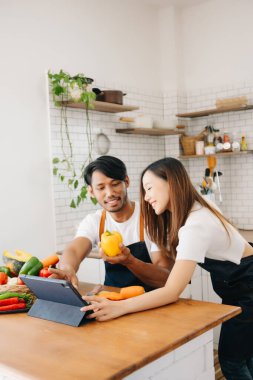 Mutlu Asyalı genç çift, ekmek ve meyveli sebzelerle birlikte ahşap mutfak masasında yemek pişiriyor.