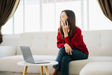 Asyalı genç bir kadın evdeki kanepede akıllı telefon ve dizüstü bilgisayar kullanıyor.