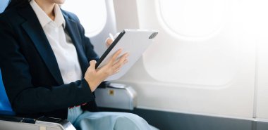 Çekici Asyalı kadın uçak yolcusu kablosuz bağlantıyı kullanarak dijital tablet üzerinde çalışırken rahat koltukta oturuyor. Stil içinde seyahat et, zarafetle çalış.