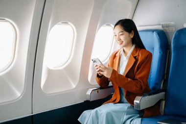 Çekici Asyalı kadın yolcu rahat koltukta oturmuş kablosuz bağlantıyı kullanarak akıllı telefon üzerinde çalışıyor. Stil içinde seyahat et, zarafetle çalış.