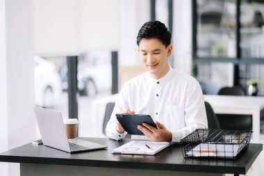 Tablet ve dizüstü bilgisayarda yazan yakışıklı genç adam ahşap masa modern ofisinde otururken.