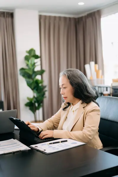 Mature businesswoman executive manager looking at  tablet, watching online webinar training or having virtual meeting video conference doing market research working in office.