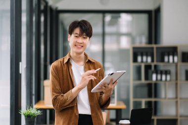 Modern ofiste elinde tablet bilgisayarla gülümseyen kendine güvenen Asyalı adam. 