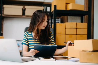 Genç işyeri sahibi kadın paket kutusu hazırlıyor ve online alışveriş konsepti üzerinde müşteriye teslim edilmesi için online olarak kontrol ediliyor.