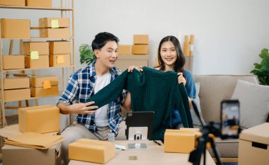 İki Asyalı blogcu, dükkanında canlı yayın yapmak için kameranın önünde kıyafetlerini gösteriyor. Çevrimiçi alışveriş arabası fikri. Evdeki kanepede.