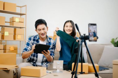 İki Asyalı blogcu, dükkanında canlı yayın yapmak için kameranın önünde kıyafetlerini gösteriyor. Çevrimiçi alışveriş arabası fikri. Evdeki kanepede.