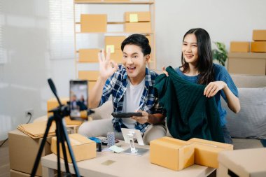 İki Asyalı blogcu, dükkanında canlı yayın yapmak için kameranın önünde kıyafetlerini gösteriyor. Çevrimiçi alışveriş arabası fikri. Evdeki kanepede.