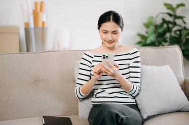 Mutlu Asyalı kadın evdeki koltukta otururken akıllı telefon kullanıyor.