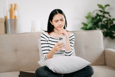 Genç Asyalı kadın rahat bir kanepede rahat rahat oturup akıllı telefonuna mesaj atıyor, gülümseyen kız evden kanepeye cep telefonu alışverişi yapıyor.