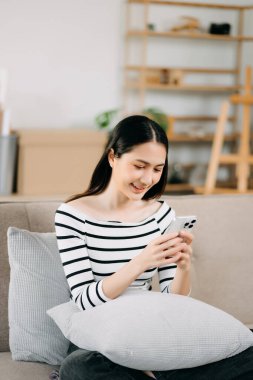 Genç Asyalı kadın rahat bir kanepede rahat rahat oturup akıllı telefondan mesajlaşıyor ve gülümseyen kız evden kanepeye cep telefonu alışverişi yapıyor.
