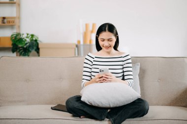 Genç Asyalı kadın rahat bir kanepede rahat rahat oturup akıllı telefonuna mesaj atıyor, gülümseyen kız evden kanepeye cep telefonu alışverişi yapıyor.