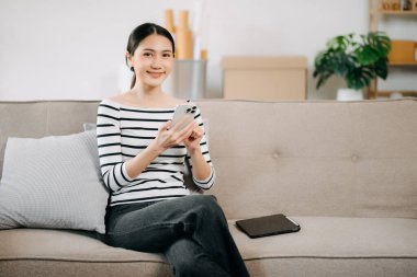 Mutlu Asyalı kadın evdeki koltukta otururken akıllı telefon kullanıyor.