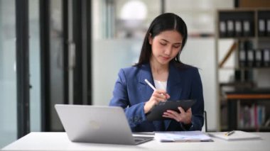 Asyalı genç iş kadını dizüstü bilgisayarla çalışıyor ve ofiste notlar alıyor.