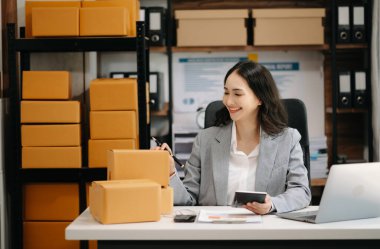 Başlangıçta küçük işletme KOBİ 'si, Girişimci sahibi Asyalı kadın dizüstü bilgisayar kullanıyor, satın alıyor ve paket ürün kutularını hazırlamak için online alışveriş siparişlerini kontrol ediyor