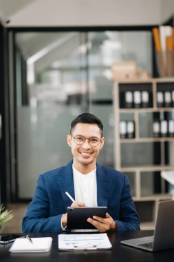 Genç ve çekici Asyalı iş adamı gülümseyip ofiste kameraya bakıyor.   