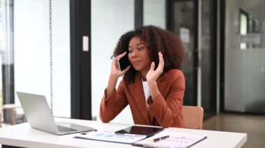 Dizüstü bilgisayarla çalışan Afrikalı iş kadını ofiste akıllı telefondan konuşuyor. 