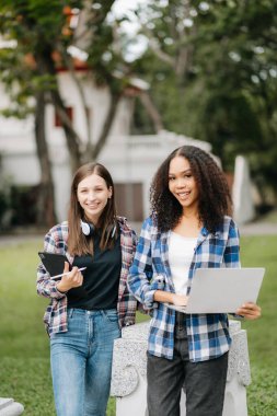 İki kız öğrenci parkta birlikte yürüyor, ellerinde tablet ve laptop var. 