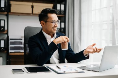 Takım elbiseli Asyalı genç bir ofis çalışanı ofiste çalışıyor ve gülümsüyor. 