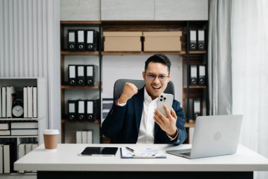 Takım elbiseli Asyalı genç bir ofis çalışanı ofiste çalışıyor ve gülümsüyor. 