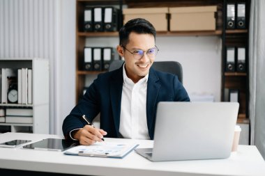 Takım elbiseli Asyalı genç bir ofis çalışanı ofiste çalışıyor ve gülümsüyor. 