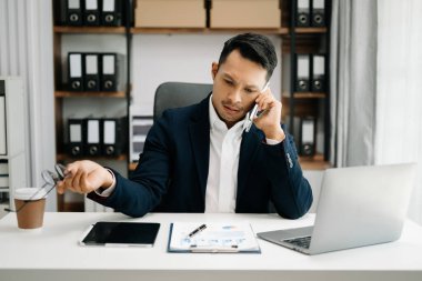 Takım elbiseli Asyalı genç bir ofis çalışanı ofiste çalışıyor ve gülümsüyor. 