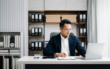 Takım elbiseli Asyalı genç bir ofis çalışanı ofiste çalışıyor ve gülümsüyor. 