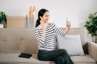 Genç Asyalı kadın evinde rahat bir kanepede rahat rahat akıllı telefondan mesaj atıyor.