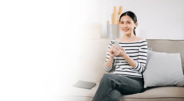 Mutlu Asyalı kadın evdeki koltukta otururken akıllı telefon kullanıyor.