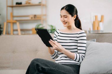 Mutlu Asyalı kadın evdeki koltukta otururken dijital tablet kullanıyor.