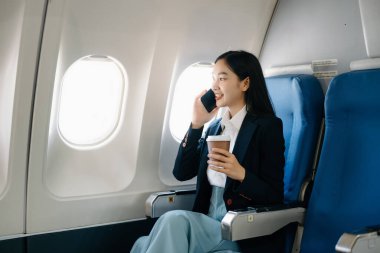 Çekici Asyalı kadın uçak yolcusu rahat koltukta oturmuş akıllı telefonla konuşuyor, kablosuz bağlantı kullanıyor. Stil içinde seyahat et, zarafetle çalış. 
