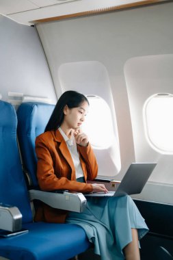 Çekici Asyalı kadın uçak yolcusu dizüstü bilgisayarda kablosuz bağlantıyla çalışırken rahat koltukta oturuyor. Stil içinde seyahat et, zarafetle çalış.