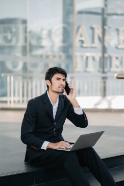 Genç Asyalı işadamı ofis binasının önünde telefonla konuşurken dizüstü bilgisayar kullanıyor. 