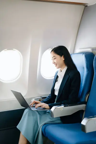 Attractive Asian female passenger of airplane sitting in comfortable seat while working laptop, using wireless connection. Travel in style, work with grace