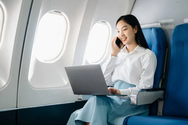 Attractive Asian female passenger of airplane sitting in comfortable seat while working on laptop computer,  using wireless connection. Travel in style, work with grace
