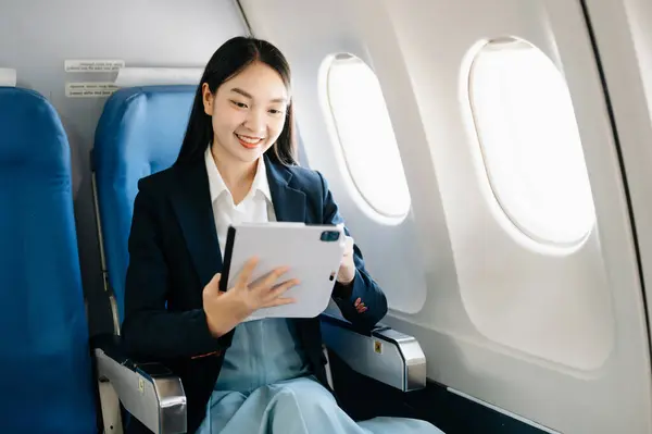 Attractive Asian female passenger of airplane sitting in comfortable seat while working on digital tablet,  using wireless connection. Travel in style, work with grace