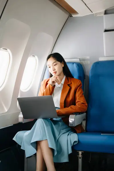 Attractive Asian female passenger of airplane sitting in comfortable seat while working on laptop computer,  using wireless connection. Travel in style, work with grace