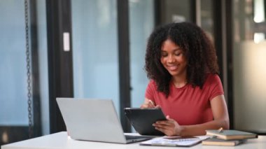 Ofiste dizüstü bilgisayarla çalışan ve tablet bilgisayarına not alan mutlu Afrikalı iş kadını. 
