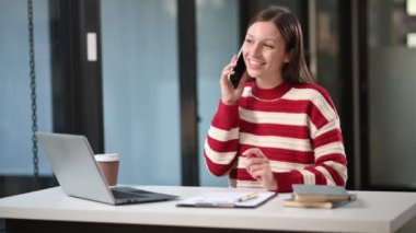Bilgisayarla çalışan ve ofiste cep telefonuyla konuşan Kafkasyalı mutlu iş kadını. 