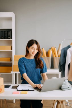 Asyalı terzi kadın terzi atölyesinde giysiler üzerinde çalışıyor. Dizüstü bilgisayarı ve stüdyosunda telefonu olan güzel bir bayan moda tasarımcısı.