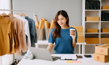 Asyalı terzi kadın terzi atölyesinde giysiler üzerinde çalışıyor. Stüdyoda dizüstü bilgisayarı, telefonu ve kahvesi olan güzel bir bayan moda tasarımcısı.
